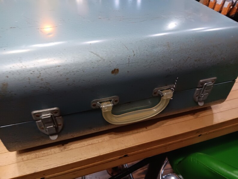 May include: A vintage, blue-green metal suitcase with a clear plastic handle and metal latches. The suitcase shows signs of wear, with scratches and a few spots of discoloration. The handle is a light yellow color.