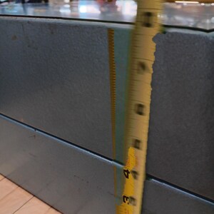 May include: A blue-grey metal cabinet with a glass top. A yellow measuring tape shows the cabinet's height is approximately 4 inches. The cabinet is in a store setting.