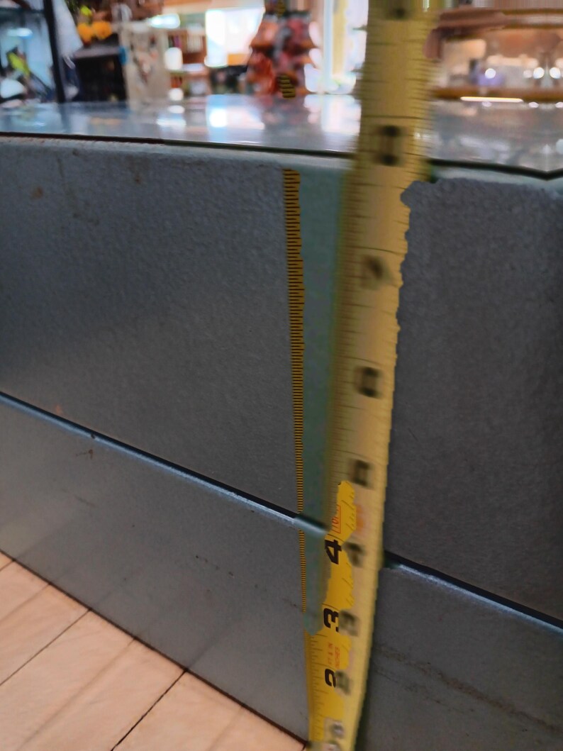 May include: A blue-grey metal cabinet with a glass top. A yellow measuring tape shows the cabinet's height is approximately 4 inches. The cabinet is in a store setting.