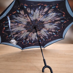 May include: A black umbrella with a feather design in shades of blue, purple, and orange. The umbrella has a light blue border and a black handle with a unique curved design. The umbrella is open, showcasing the intricate pattern.