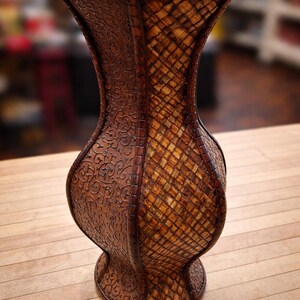 May include: A decorative brown vase with a unique hourglass shape. The vase features intricate embossed floral patterns on one side and a woven, grid-like design on the other. The vase is made of a dark brown material.