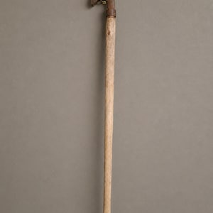 May include: A vintage walking stick with a curved metal handle and a wooden shaft. The handle is a dark, aged metal, possibly brass or bronze, and is attached to the wooden shaft with a metal band. The wood is a light, natural color.