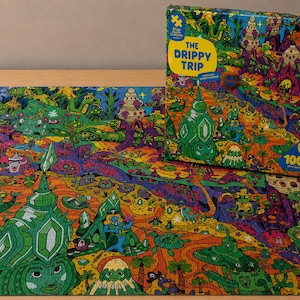 May include: A 1000-piece jigsaw puzzle titled The Drippy Trip by The Magic Puzzle Company. The puzzle and its box feature a vibrant, psychedelic illustration with green, orange, and purple colors, depicting surreal landscapes, creatures, and structures.