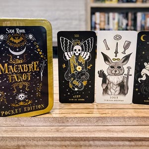 May include: A tarot card set with a gold-colored tin featuring the text "The Macabre Tarot Pocket Edition." Three tarot cards are displayed, each with unique illustrations in black, white, and gold. The cards feature images of a moth, a rabbit, and a crow.