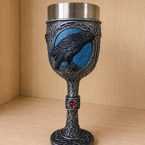 May include: A decorative goblet with a silver finish, featuring a detailed raven design. The raven is set against a blue background within a circular frame. A red jewel accents the stem, and the base has an intricate Celtic knot pattern.
