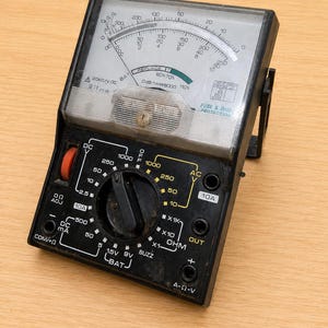 May include: A black analog multimeter featuring a large white display with multiple measurement scales, a central rotary dial for selecting settings, and various input ports labeled for DC V, AC V, DC mA, and Ohm measurements with clear text.