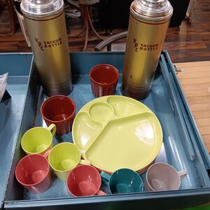 May include: A vintage picnic set in a blue metal case. Includes two gold vacuum bottles with cork stoppers, a yellow divided plate, and several colorful cups in red, green, teal, and gray. The bottles have the text "VACUUM BOTTLE".