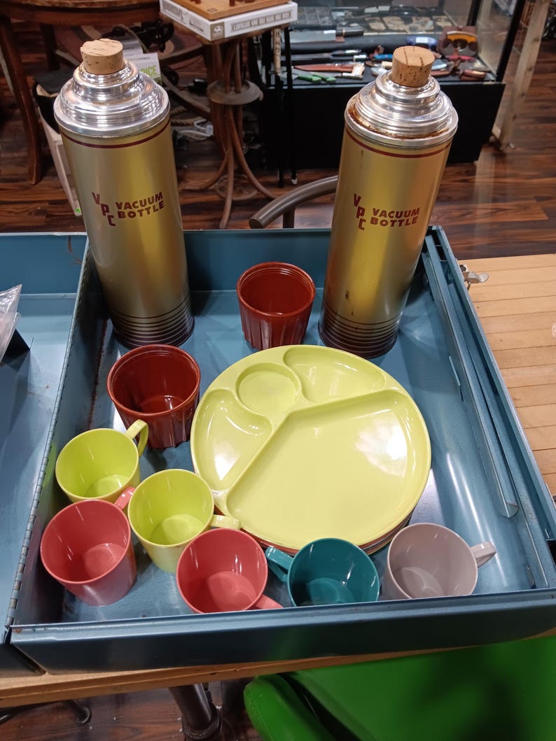 May include: A vintage picnic set in a blue metal case. Includes two gold vacuum bottles with cork stoppers, a yellow divided plate, and several colorful cups in red, green, teal, and gray. The bottles have the text "VACUUM BOTTLE".