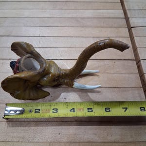 May include: A ceramic elephant head figurine, painted in shades of brown and tan, with white tusks. The trunk curves upwards. The figurine is approximately 5 inches long, as measured by a yellow tape measure.