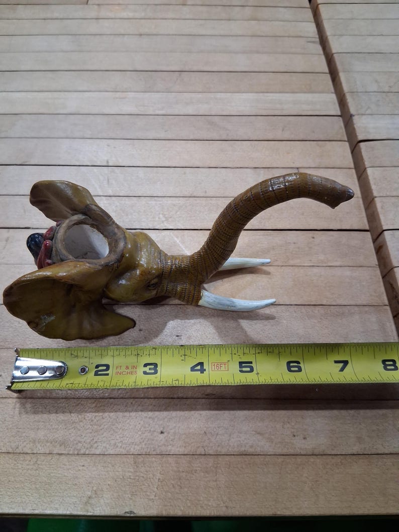 May include: A ceramic elephant head figurine, painted in shades of brown and tan, with white tusks. The trunk curves upwards. The figurine is approximately 5 inches long, as measured by a yellow tape measure.