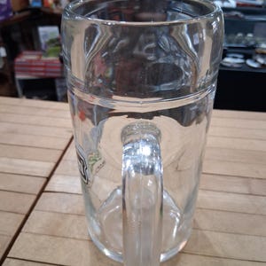May include: A clear glass beer mug featuring a sturdy handle and a smooth, cylindrical body. The mug has a thick base and a wide rim, with a partial logo visible on the side. The design is simple and functional, crafted from transparent glass.