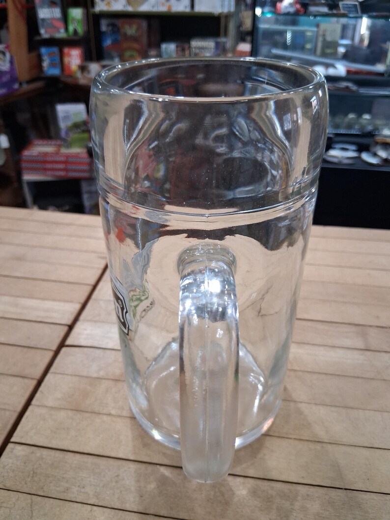 May include: A clear glass beer mug featuring a sturdy handle and a smooth, cylindrical body. The mug has a thick base and a wide rim, with a partial logo visible on the side. The design is simple and functional, crafted from transparent glass.
