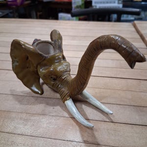 May include: A detailed, handcrafted elephant head sculpture with a trunk and tusks. The sculpture is primarily brown with lighter accents on the tusks and ears. The trunk curves gracefully, and the tusks are white. The elephant head is a decorative item.