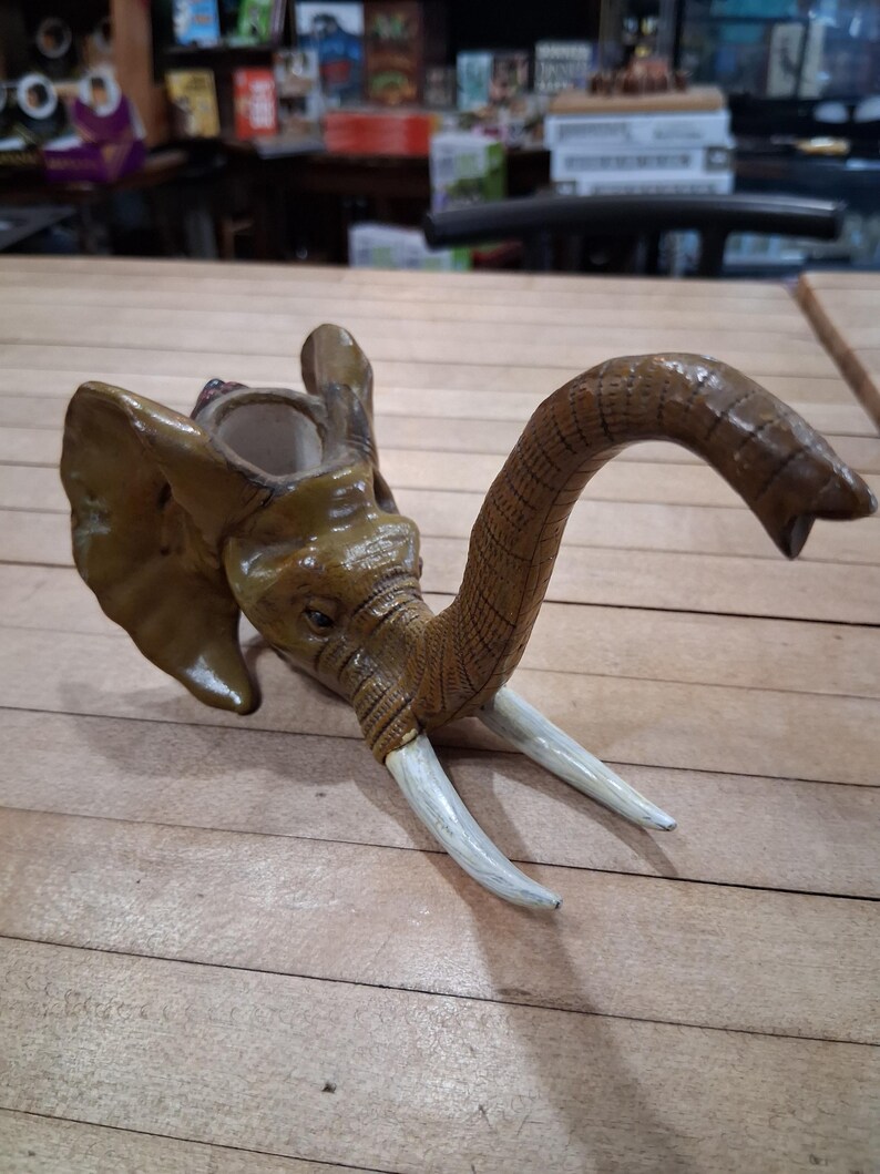May include: A detailed, handcrafted elephant head sculpture with a trunk and tusks. The sculpture is primarily brown with lighter accents on the tusks and ears. The trunk curves gracefully, and the tusks are white. The elephant head is a decorative item.