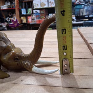 May include: A detailed, brown elephant figurine with ivory-colored tusks. The elephant's trunk is curved upwards. The figurine is approximately 2 inches tall, as measured by a yellow tape measure.