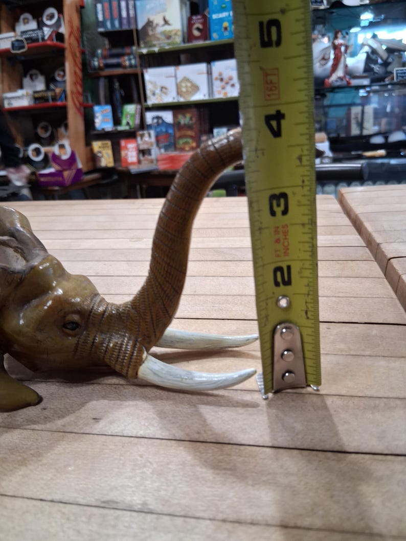 May include: A detailed, brown elephant figurine with ivory-colored tusks. The elephant's trunk is curved upwards. The figurine is approximately 2 inches tall, as measured by a yellow tape measure.