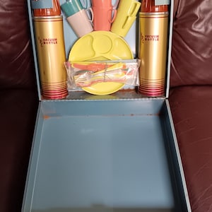 May include: Vintage picnic set in a blue metal case. Includes two gold vacuum bottles with red accents, colorful cups, plates, and cutlery. The vacuum bottles have the text "VACUUM BOTTLE".