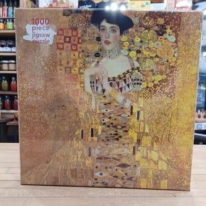 May include: A 1000-piece jigsaw puzzle featuring a portrait of a woman in a gold dress. The artwork has a gold and brown color scheme with intricate patterns and details. The puzzle box has the text "1000 piece jigsaw puzzle".