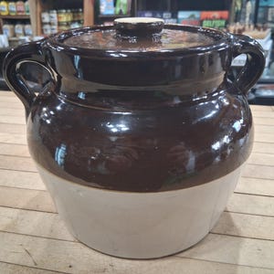 May include: A two-tone ceramic crock pot with a lid. The upper portion is a glossy dark brown, while the lower section is a creamy white. It has two handles and a small knob on the lid. This kitchenware is suitable for cooking and storage.