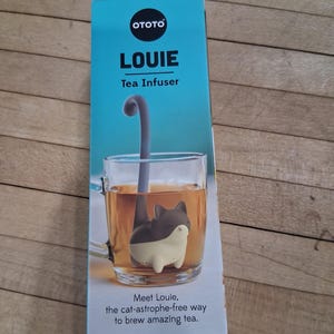 May include: A teal box with the word "LOUIE" and "Tea Infuser" printed on it. Inside a clear glass mug is a cat-shaped tea infuser. The infuser is gray and white, with a long gray handle. The mug contains tea.
