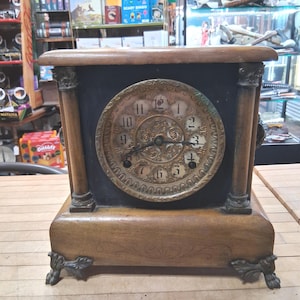 May include: An antique wooden clock with a round, ornate face featuring Roman numerals. The clock has a dark face, a wooden frame, and decorative metal feet. The clock's face is gold-toned with black hands.