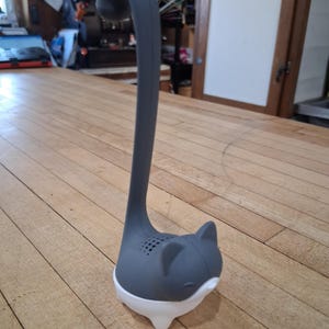 May include: A gray and white cat-shaped tea infuser. The infuser has a long, curved handle and a cat face design with small holes for steeping tea. The base is white, and the cat's head and handle are gray.