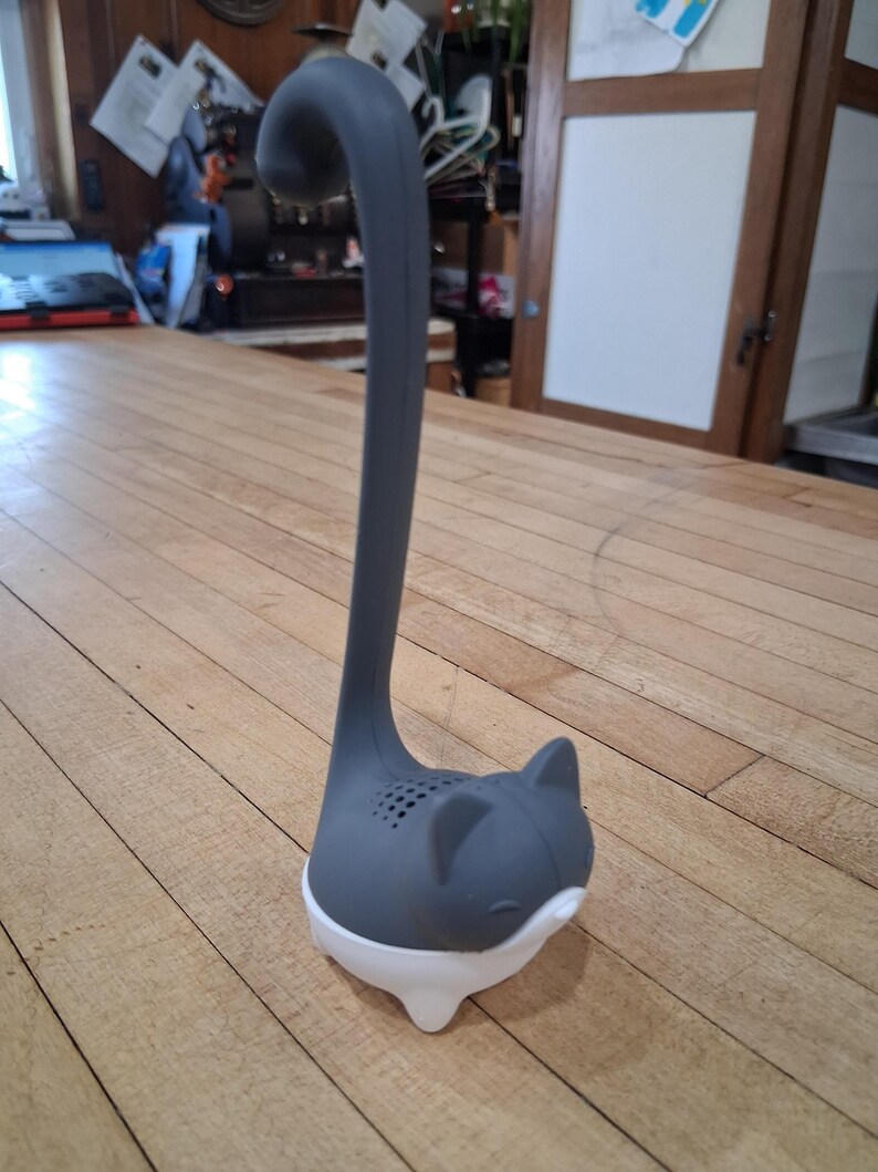May include: A gray and white cat-shaped tea infuser. The infuser has a long, curved handle and a cat face design with small holes for steeping tea. The base is white, and the cat's head and handle are gray.