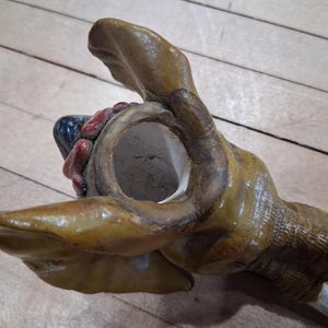 May include: A detailed, close-up view of a ceramic dog head, likely a decorative item. The dog's head is painted in shades of brown and tan, with visible details of the snout, mouth, and ears. The interior of the mouth is red and black.