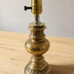 May include: A brass lamp base with a rounded, tiered body featuring embossed arch patterns. The top includes a metallic socket with a black turn knob and visible text markings. The structure is polished brass with a reflective surface finish.