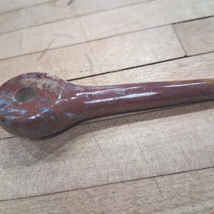 May include: A reddish-brown, ceramic pipe with a rounded bowl and a long, cylindrical stem. The pipe has a glossy finish with blue and white accents. The bowl features a circular hole. The object is resting on a wooden surface.