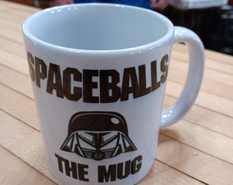Könnte beinhalten: Weiße Keramik-Tasse mit den Worten "SPACEBALLS" und "THE MUG" in dunkelbraunen Buchstaben. Eine dunkelbraune Helm-Grafik befindet sich unter dem Wort "SPACEBALLS". Die Tasse hat einen gebogenen Henkel und steht auf einer hellbraunen Holzoberfläche.