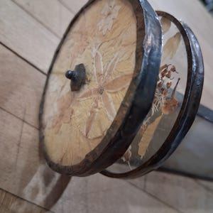 May include: A decorative, handcrafted object featuring two circular, wooden frames with dried flowers and leaves encased in clear material. The frames are connected by a dark metal handle and rimmed with a dark, textured metal.