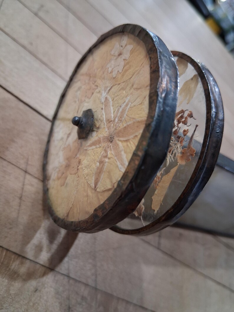 May include: A decorative, handcrafted object featuring two circular, wooden frames with dried flowers and leaves encased in clear material. The frames are connected by a dark metal handle and rimmed with a dark, textured metal.