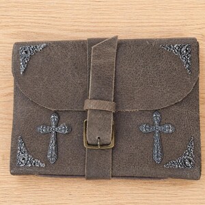 May include: A brown leather clutch with a buckle closure. The clutch features decorative metal crosses and corner accents. The leather has a textured appearance.