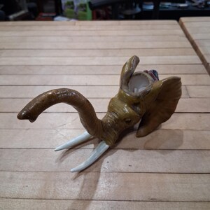 May include: A brown elephant head sculpture with white tusks. The elephant's trunk is curved, and its ears are flared. The sculpture is made of a light brown material and sits on a wooden surface.