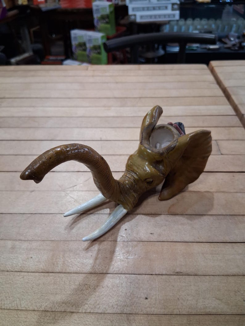 May include: A brown elephant head sculpture with white tusks. The elephant's trunk is curved, and its ears are flared. The sculpture is made of a light brown material and sits on a wooden surface.