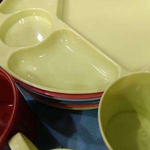 May include: A stack of colorful plastic plates and cups. The top plate is divided into three sections and is a pale yellow color. Other plates are red, teal, and pink. A red cup and a yellow cup are also visible.