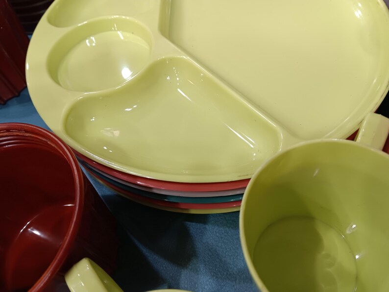 May include: A stack of colorful plastic plates and cups. The top plate is divided into three sections and is a pale yellow color. Other plates are red, teal, and pink. A red cup and a yellow cup are also visible.