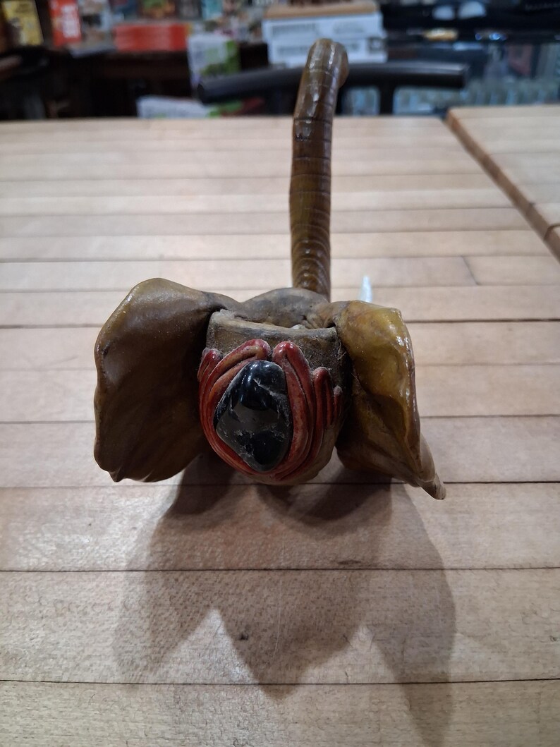 May include: A handcrafted, brown elephant sculpture with large, textured ears and a long, curved trunk. The elephant's face features a dark, glossy stone and red accents, creating a unique and artistic piece.