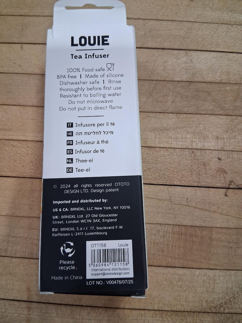 May include: A white and black box for a tea infuser, with the name "LOUIE" at the top. The box includes text about the product's features, such as being food safe and dishwasher safe. The box also has a recycling symbol.