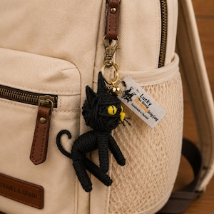 May include: A black crocheted cat keychain with yellow eyes and whiskers hangs from a beige backpack. The backpack has brown leather accents and a mesh side pocket. A small tag with text is attached to the keychain.