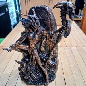 May include: Bronze-toned statue of a female figure with horns, holding a staff topped with a skull. The statue has intricate details, including a serpent-like design on the staff and a base with skulls and bones. The Divinity Collection is printed on the box in the background.