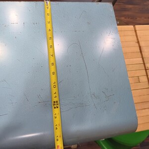 May include: A light blue, metal table with a rounded edge. A yellow measuring tape shows the table is approximately 17 inches long. The table has visible wear and tear. A green chair is partially visible.