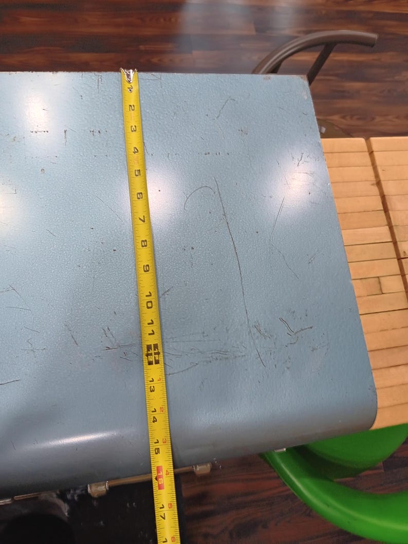 May include: A light blue, metal table with a rounded edge. A yellow measuring tape shows the table is approximately 17 inches long. The table has visible wear and tear. A green chair is partially visible.