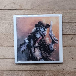 May include: Square ceramic tile featuring a detailed illustration of a zombie cowboy holding a revolver. The artwork depicts the undead figure in a black hat and coat, with a red neckerchief, set against a gradient orange and brown background.