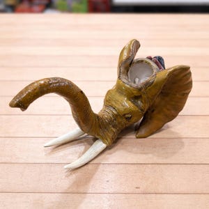 May include: A handcrafted, tan-colored elephant figurine with white tusks. The elephant's trunk is curved, and its head is tilted upwards. The figurine is made of a textured material and sits on a light brown wooden surface.