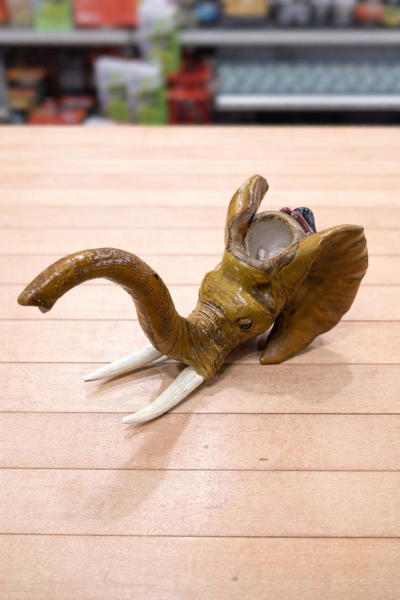 May include: A handcrafted, tan-colored elephant figurine with white tusks. The elephant's trunk is curved, and its head is tilted upwards. The figurine is made of a textured material and sits on a light brown wooden surface.