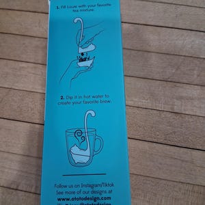 May include: A teal box with instructions for using a tea infuser. The box features illustrations of a tea infuser being filled and steeped in a mug. Text on the box includes "Fill Louie with your favorite tea mixture" and "Dip it in hot water to create your favorite brew."