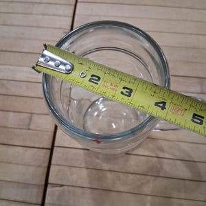 May include: A clear glass beer mug with a handle, viewed from above. A yellow measuring tape is placed across the top opening, showing a diameter of approximately 4 inches. The mug has a smooth, transparent surface and a thick, sturdy base.