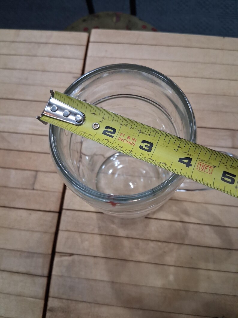 May include: A clear glass beer mug with a handle, viewed from above. A yellow measuring tape is placed across the top opening, showing a diameter of approximately 4 inches. The mug has a smooth, transparent surface and a thick, sturdy base.